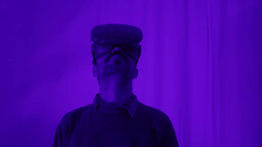 A man looks up while standing and using a virtual reality headset in a white room lit with a purple light