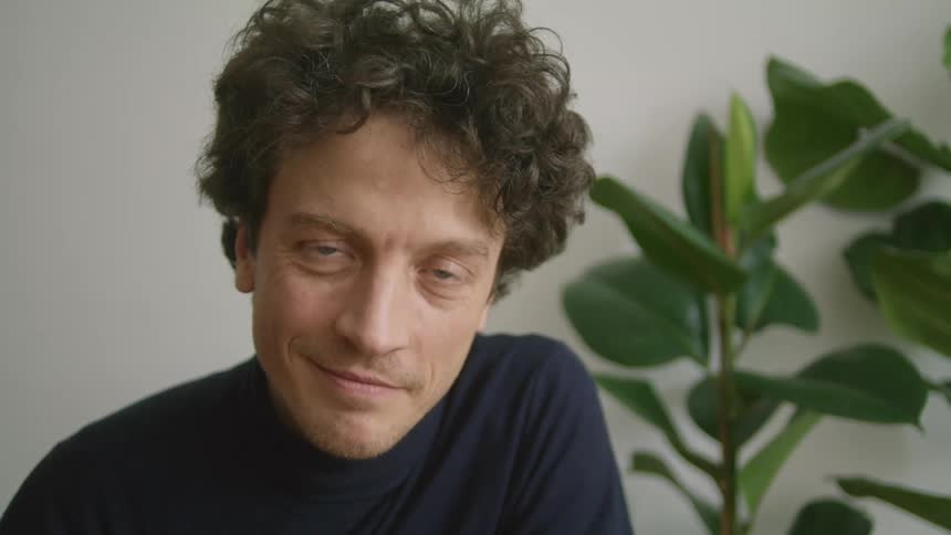 A man with curly brown hair smiling in front of a potted houseplant