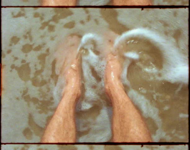 An ocean wave washes over the feet of a man standing on a beach during the day