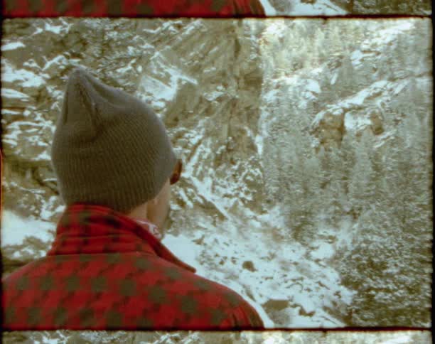 A man in warm clothing stands outdoors while looking a towering mountain range covered in snow