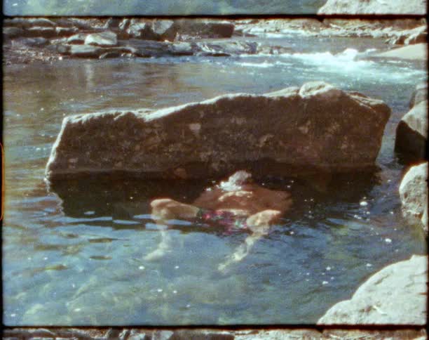 A man sits in the water as a child lays on the rocks