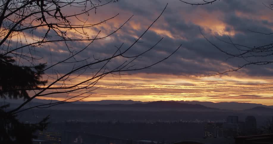 Dark clouds move over a colored sky above a city with mountains on the horizon