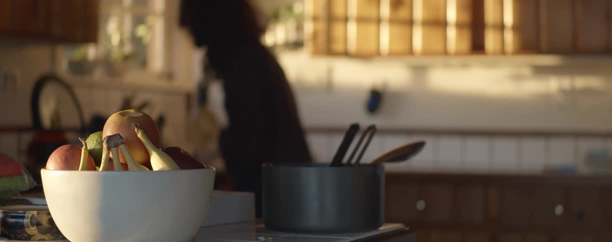 Bowl of fruit stands on kitchen counter in sunlight while silhouette of person works in kitchen