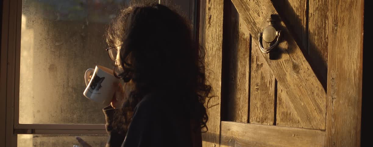 A woman walks up to her front door and looks outside while drinking from a mug