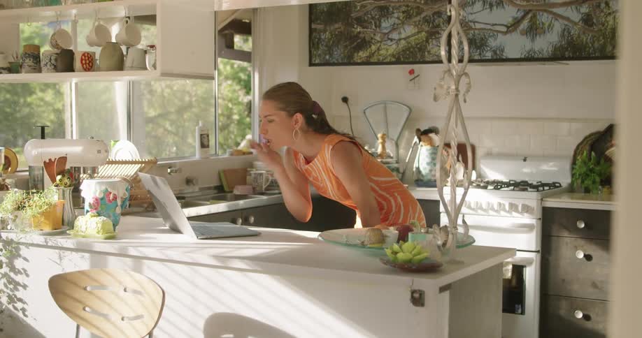 Woman wearing dress leans over kitchen counter waving and blowing kiss at laptop