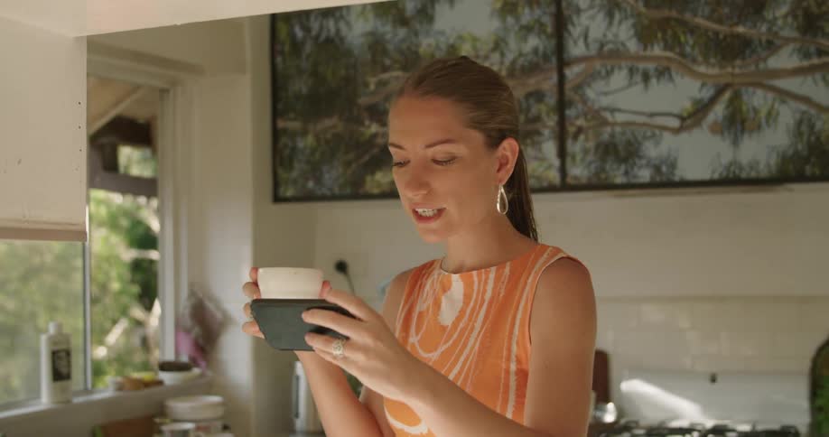A woman holds a white mug in one hand and a smartphone on the other as she stands in her kitchen and talks via video call