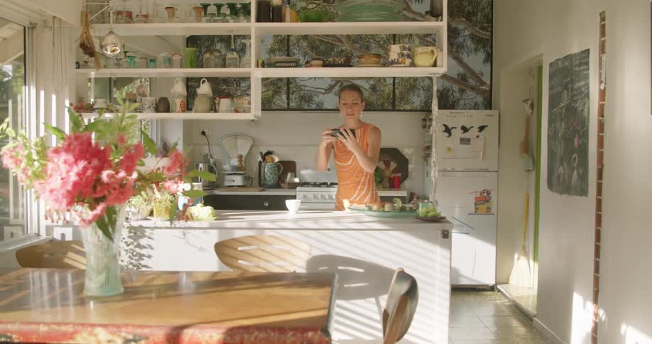 A woman stands in her kitchen and drinks coffee and waves at someone she is video chatting with on her cell phone