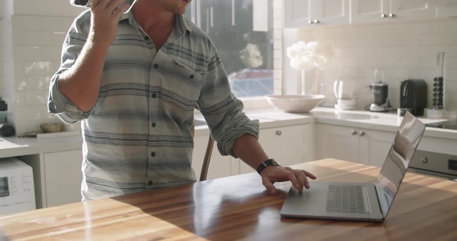 Man standing in kitchen talking on mobile phone checks laptop
