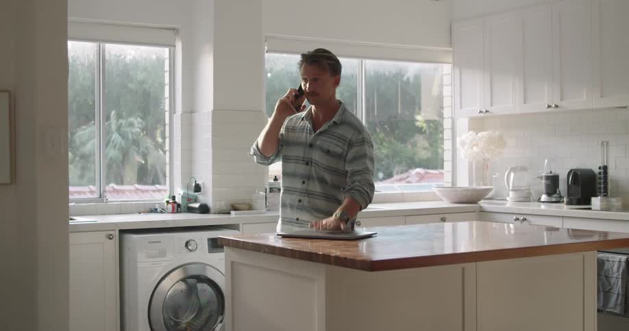 A man stands in his kitchen and talks on his cell phone