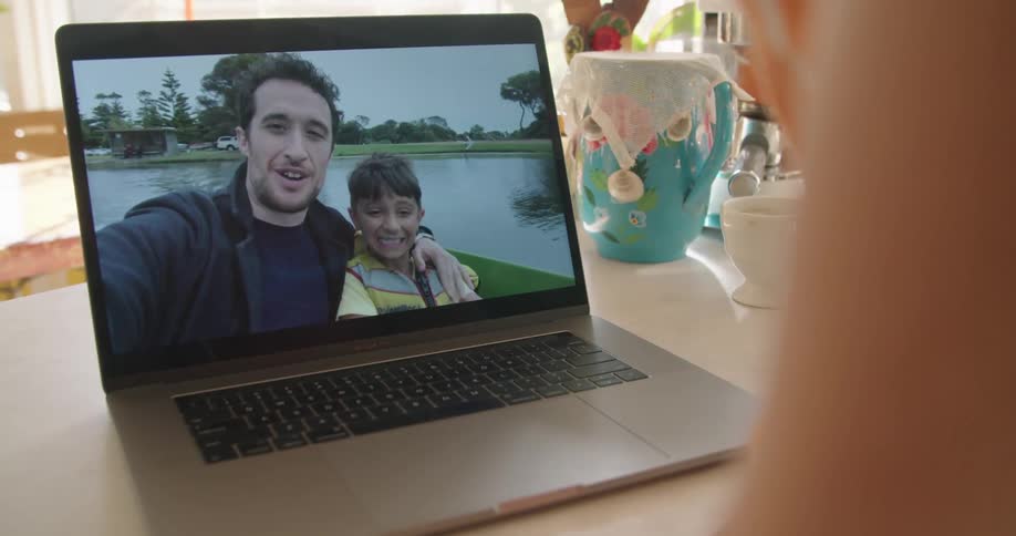 A woman stands in her kitchen and talks with her son and husband on a laptop while they are riding in a boat