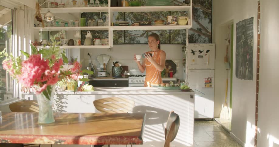 A woman holds a mug in one hand and a smartphone in the other as she talks in her kitchen during a video call