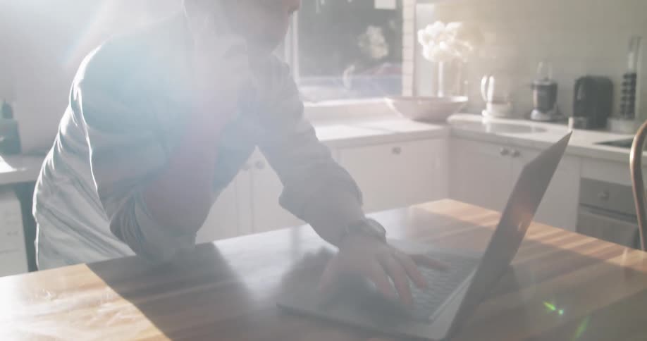 Man leaning over kitchen island types on laptop while talking on mobile phone