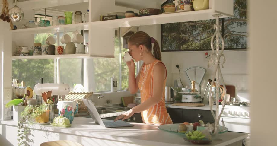 A woman standing in a kitchen typing on a laptop on a counter picks up a mug and takes a sip at daytime