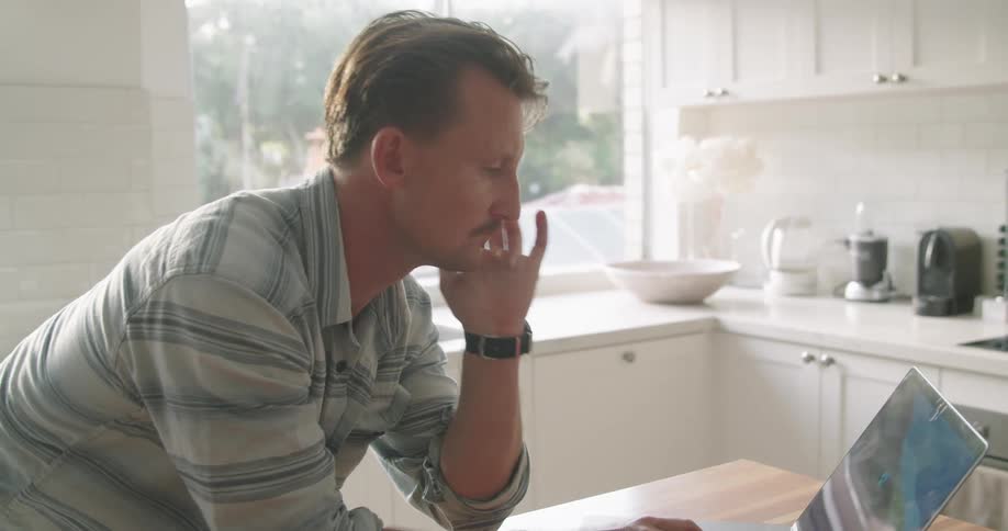 A man stands in his kitchen and types on his laptop