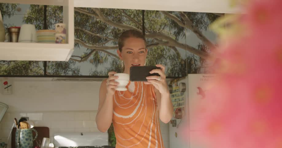 A woman drinks from a mug and sets it down to wave during a video call on her smartphone