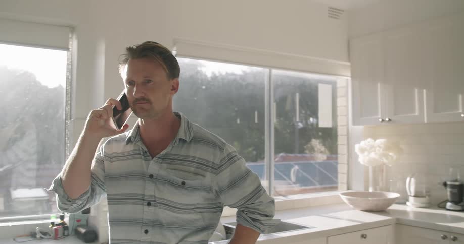 Man stands in sunny white kitchen talking on mobile phone