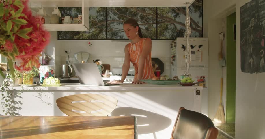 A woman stands in her kitchen and waves at someone on her laptop