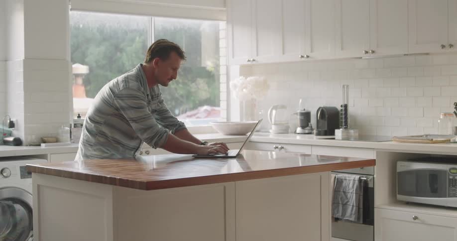 A man typing on a laptop while leaning over a kitchen counter at home