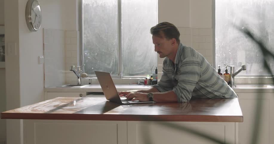 A man in a kitchen typing on a laptop on a counter with windows in the background