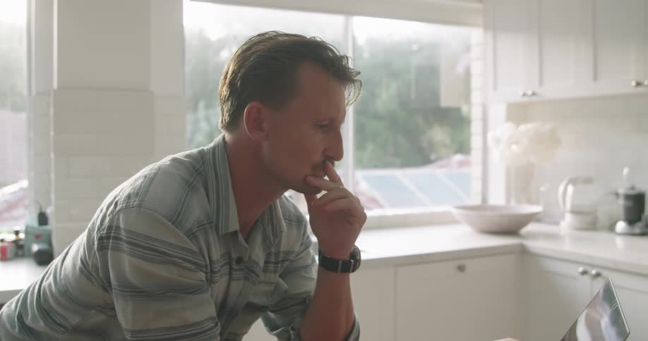A man typing on a laptop while leaning over a kitchen counter at home