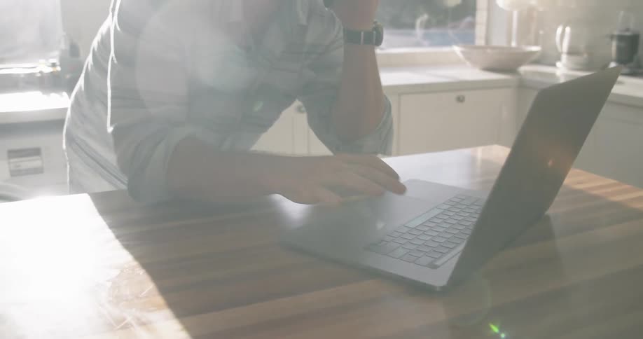 A man stands in his kitchen and talks on his cell phone while using his laptop