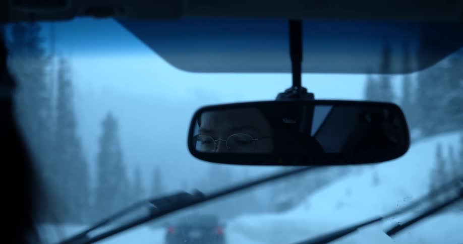 Woman drives car along snow covered road past forest topped hill following another car