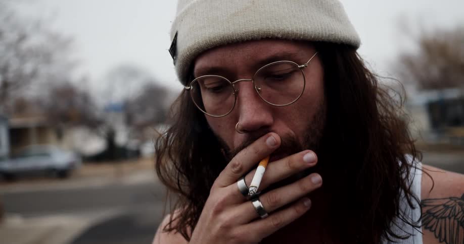 A man with long curly hair stands outside his neighborhood and smokes a cigarette