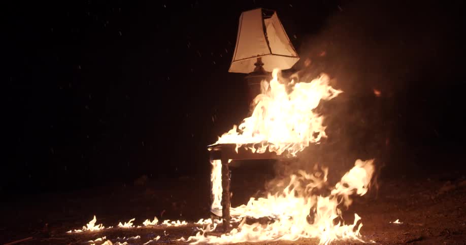 A lamp on and end table burns as flames destroy it outside at night