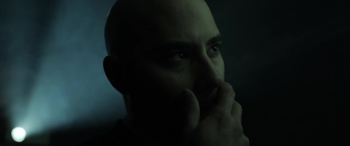 A man rubs his chin as he sits and watches with a projector light beam shining behind him