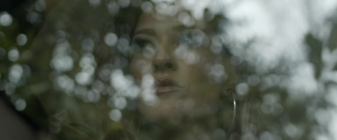 A womans face is reflected in glass as she stands outside surrounded by trees on a windy day