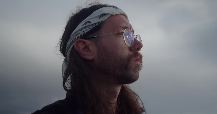 A man with a bandanna around his head and glasses with the sky and clouds in the background