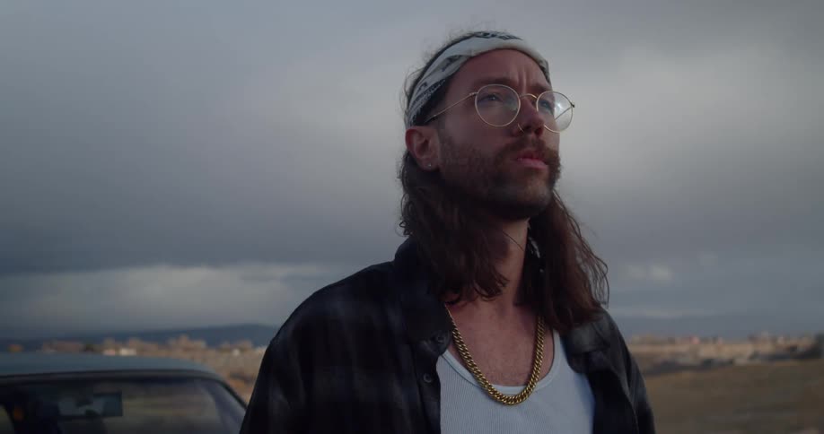 A man with glasses and a headband with a vehicle the landscape the sky and clouds in the background