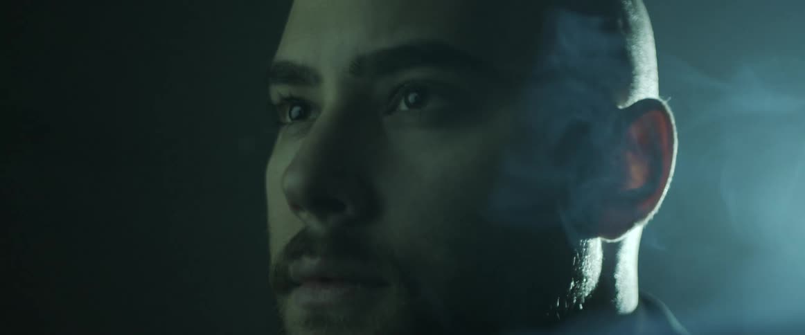 A bald man stands outside as a bright light shines on the back of his head and smoke billows around him