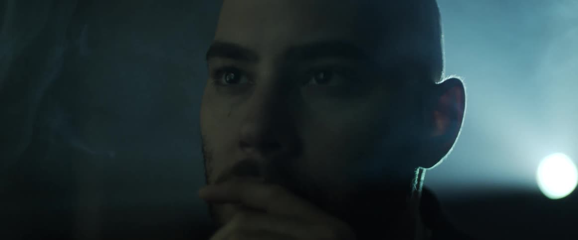 A man stands with a finger over his mouth as a bright light shines on his head and smoke fills the air around him