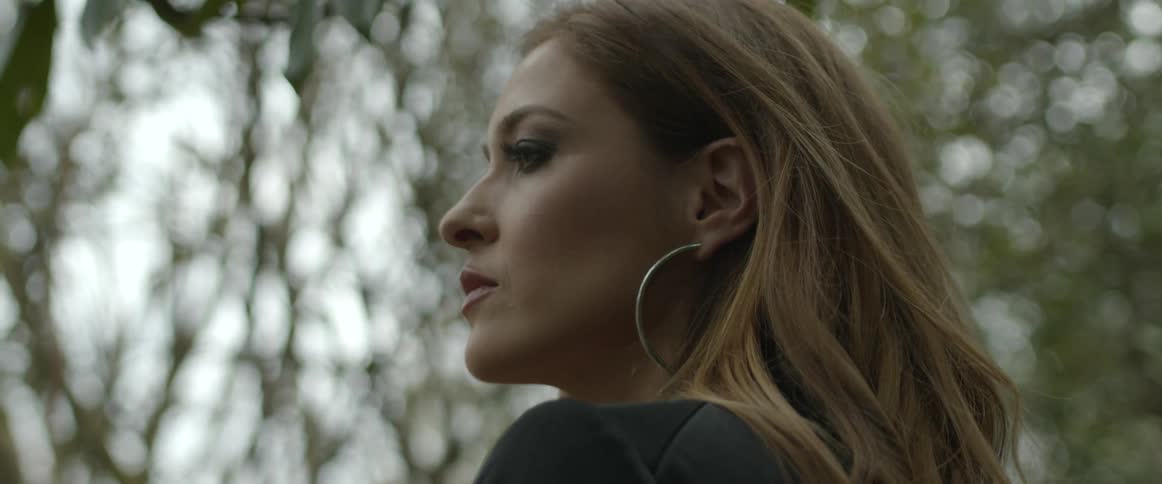 A woman flips her long brown hair behind her shoulder as she stands in a forest