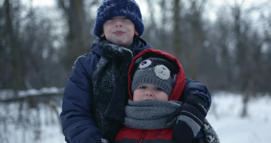 A boy holds onto his younger brother as they stand bundled up for winter in the snow