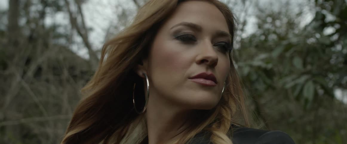 A woman in a blazer and large hoop earrings stares into the distance while standing in the woods