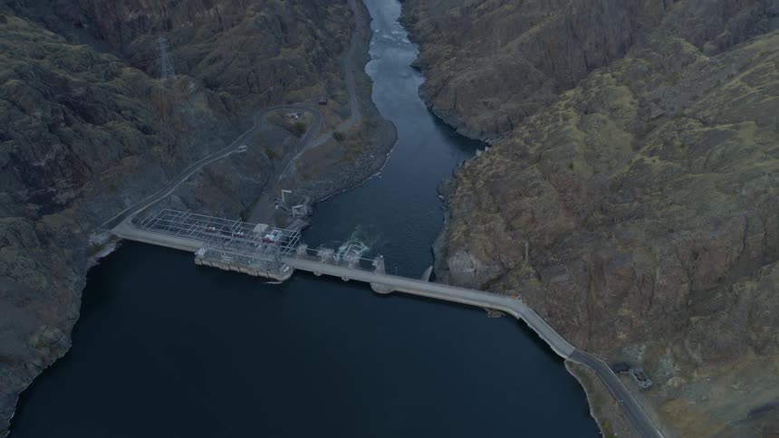Pull back tilt up reveal of a river extending away from a concrete dam built into a steep mountain valley
