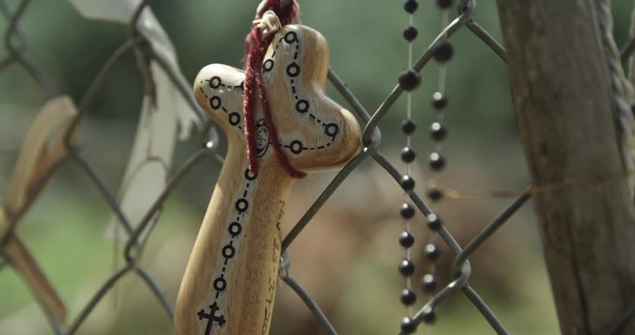 A small cross and rosary hang from a chain link fence