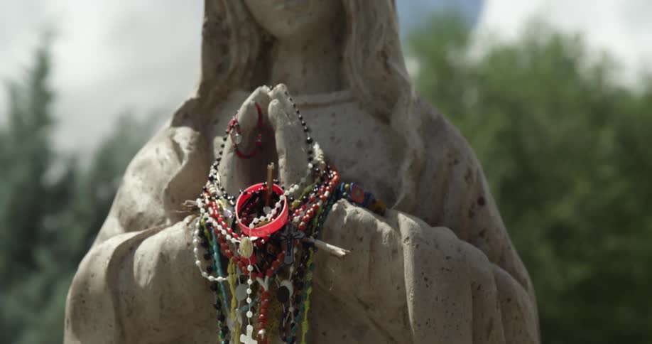Rosaries hang off religious icon standing outside under cloudy blue sky