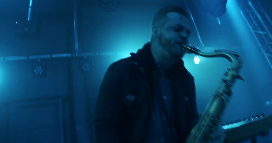 A man on a stage playing the saxophone with a keyboard next to him and flashing lights in the background