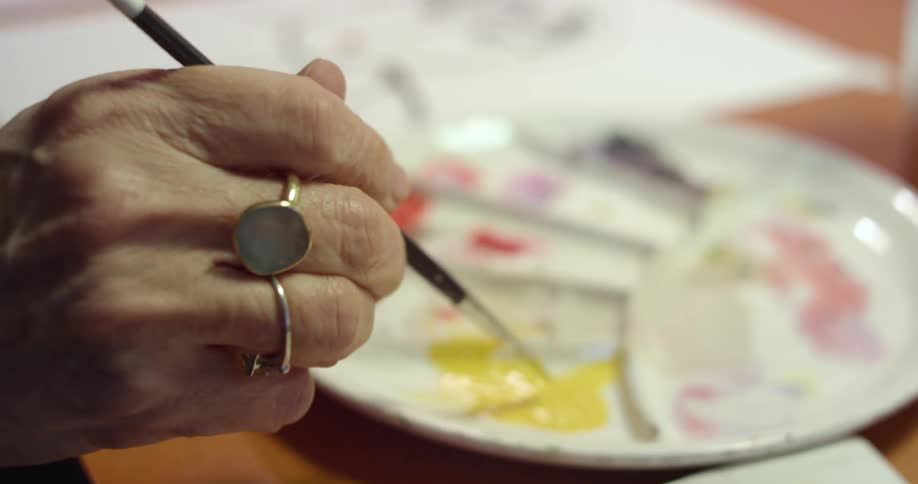 An elderly woman rubs a paintbrush in paint