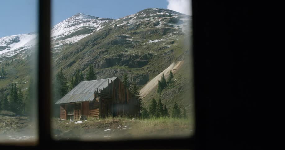A small log cabin with damaged walls sits at the bottom of a mountain