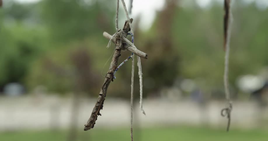 A cross made out of sticks hangs from a tree lightly swinging from strings