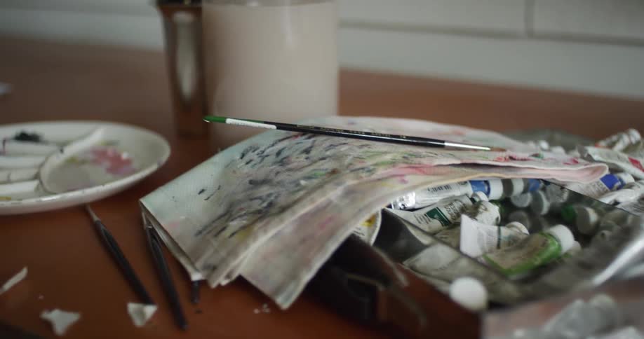 Used art supplies such as paint tubes paintbrushes and paper are shown on a table