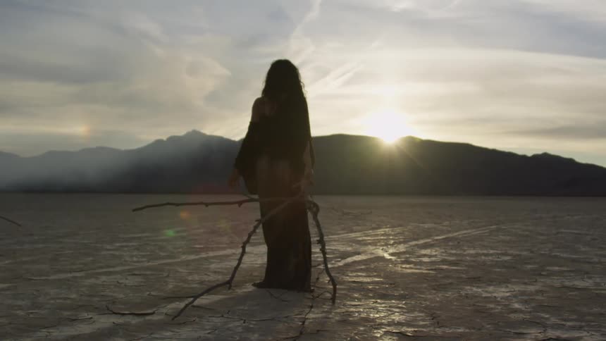 Woman dragging branch walks across plain toward sun rising over mountain