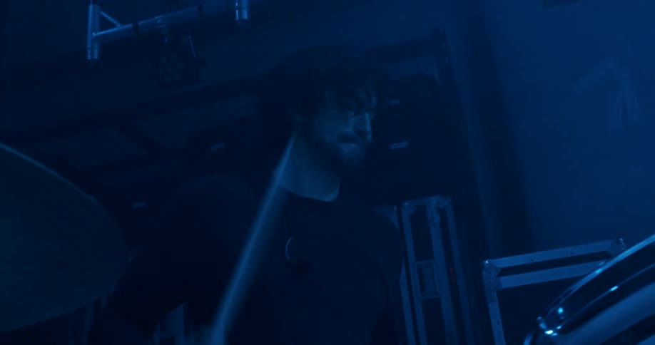 A man plays a set of drums with drumsticks inside of a dark room with a spotlight moving overhead