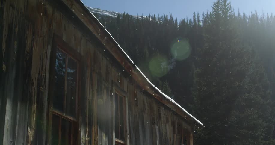 Melting snow falls from the roof of a small cabin next to a forest