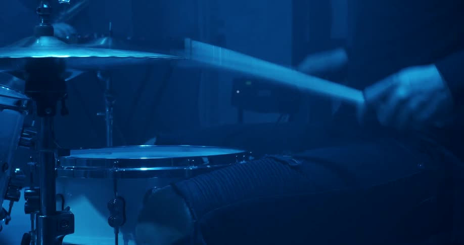 A drummer plays on a drum set in a dark blue light glow
