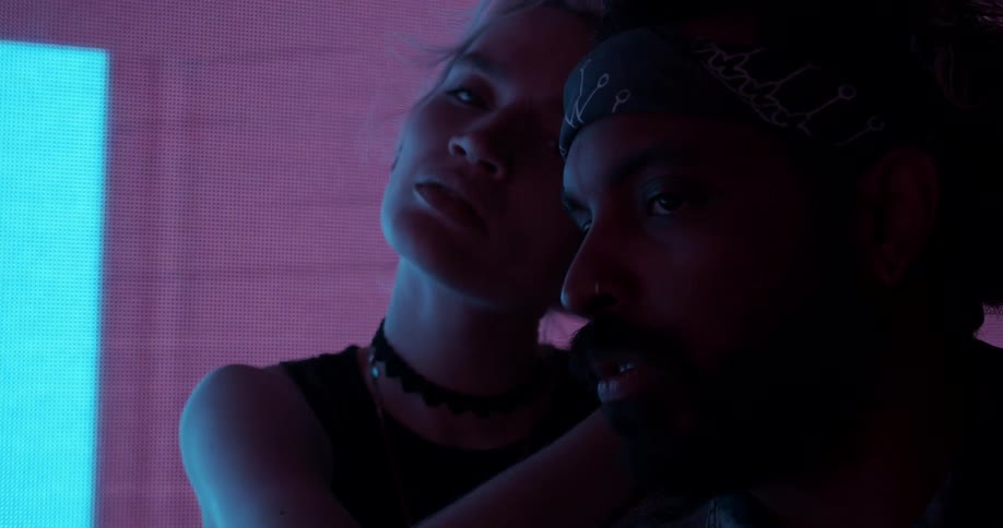 A woman poses her head and stares at the camera while holding onto a man who turns his eyes to stare at her in front of a digital screen changing colors